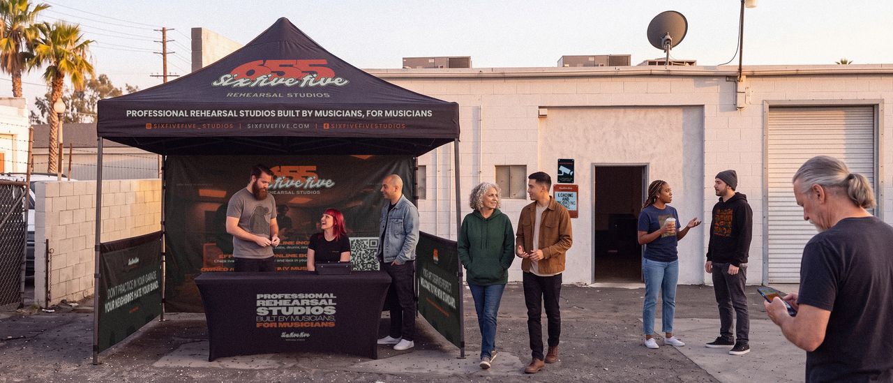 Open house at 655 Rehearsal Studios: a branded canopy and skirted table in the back lot, two staff behind the table and six visitors standing outside the studio entrance.