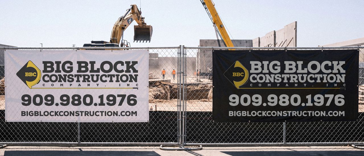 Two Big Block Construction banners on the fence of an active job site, one white and one black, with an excavator and crew working behind them.