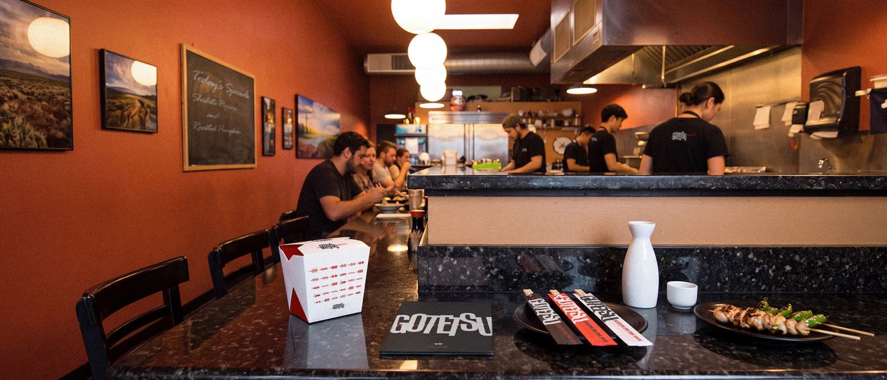 The Gotetsu dining room during service: diners at the counter, cooks working the open kitchen, and a branded takeout box, menu and chopstick sleeves in the foreground.
