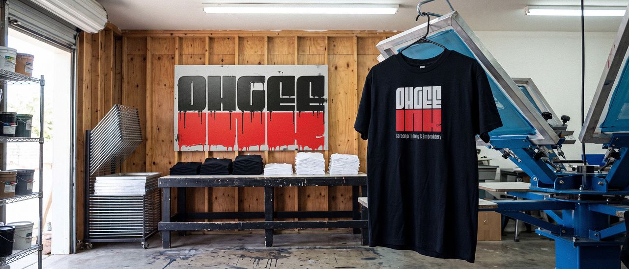 The OhGee Ink shop: the logo painted across the back wall, folded shirts on a worktable, screens racked at the left and a screen printing carousel at the right.