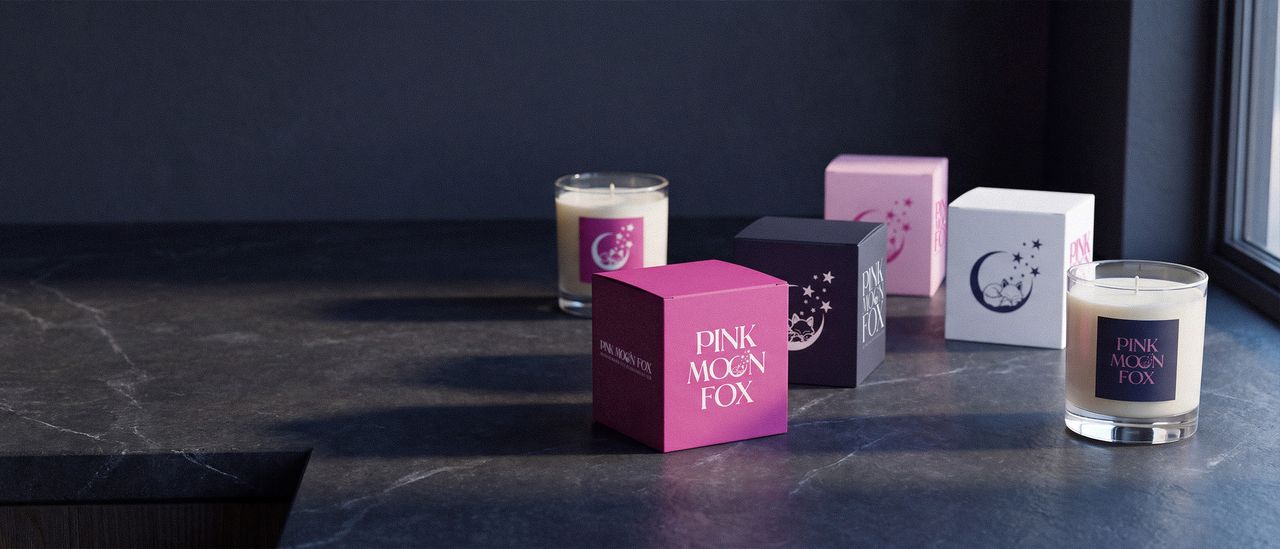 Pink Moon Fox candles and packaging on a dark stone counter: five boxes in pink, magenta, navy and white beside two glass candles in window light.