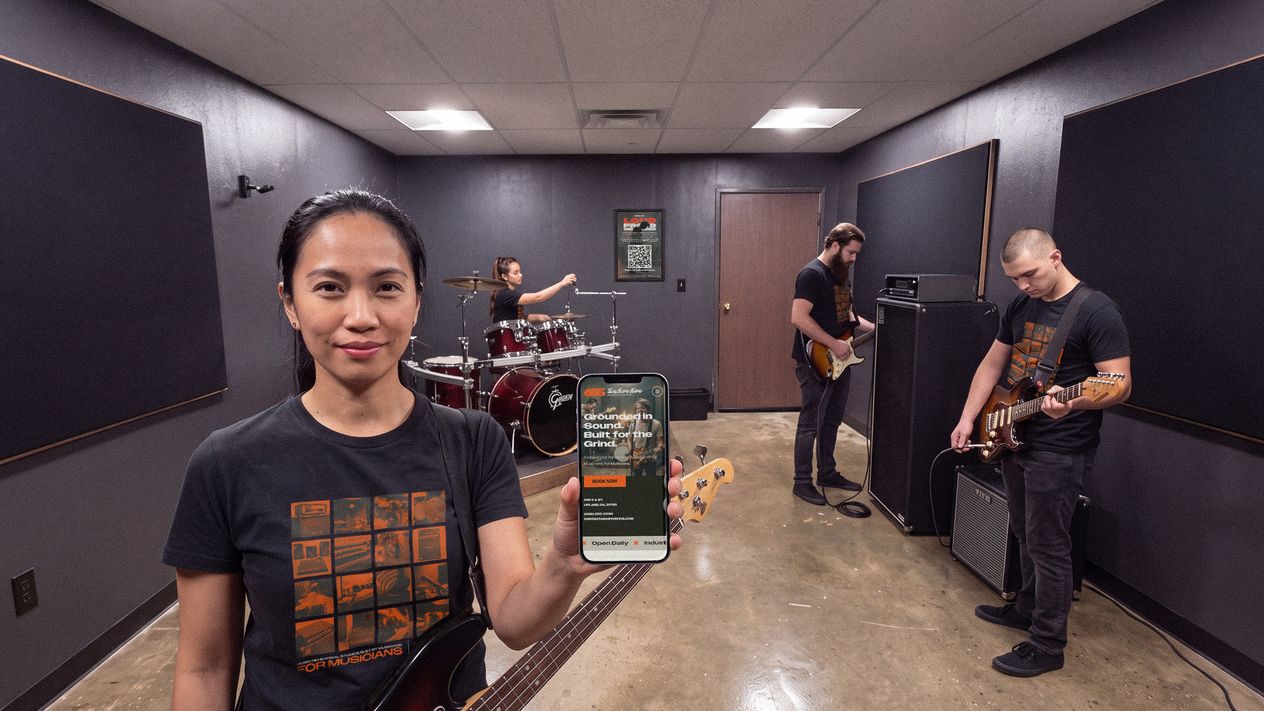 A bassist in a 655 rehearsal room holding up a phone showing the studio website, her bandmates setting up drums and guitars behind her.