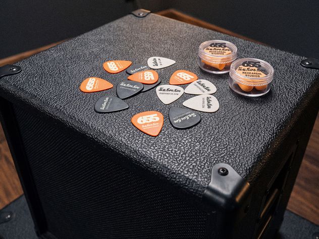 655 guitar picks in orange, black and bone scattered across the top of an amplifier cabinet, next to two branded earplug cases.