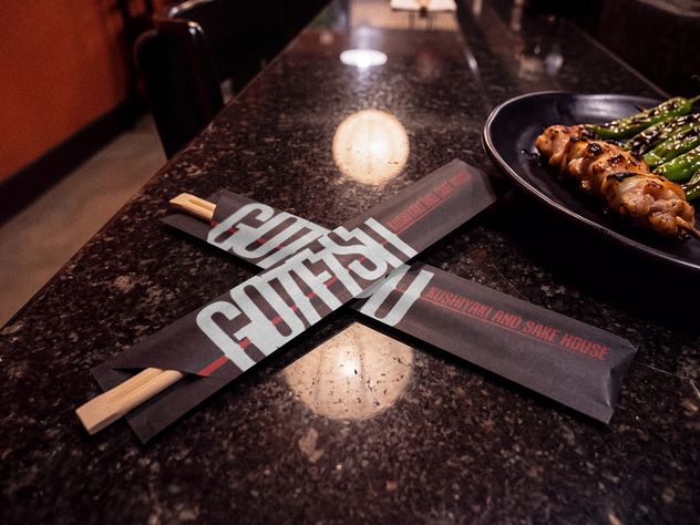 Two Gotetsu chopstick sleeves crossed on the stone counter, reading Kushiyaki and Sake House, with a plate of skewers and blistered shishito beside them.