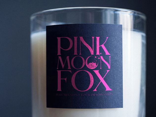 Close on a Pink Moon Fox candle label: magenta foil type on navy stock, with the line Illuminating your imagination beneath the mark.
