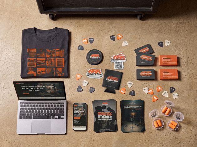 The full 655 kit laid out on a studio floor: a laptop and phone showing the website, a printed tee, stickers, a koozie, business cards, flyers, guitar picks and earplugs.