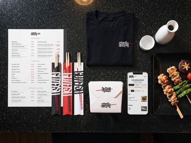 The Gotetsu system laid out on the counter: printed menu, folded tee, chopstick sleeves in three colors, takeout box, a phone showing online ordering, a sake set and a plate of skewers.