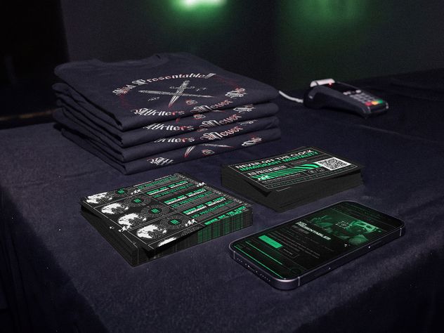A Kid Presentable!!! merch table at a show: stacked shirts, two decks of printed cards, a card reader and a phone showing the website.