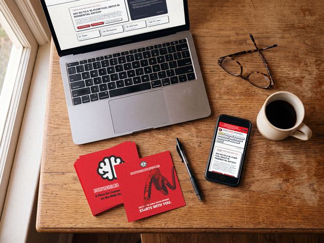 A laptop, phone and stack of red Saticoy Sensible Development cards on a wooden table beside reading glasses and a cup of coffee.