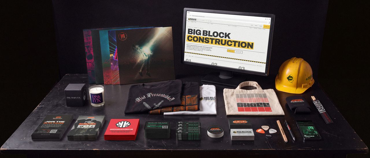 A black studio table laid out with finished work from seven different clients: record sleeves, folded printed shirts, a candle and its box, a canvas tote, a branded hard hat, stacks of flyers, stickers and business cards, and a monitor showing a client website.