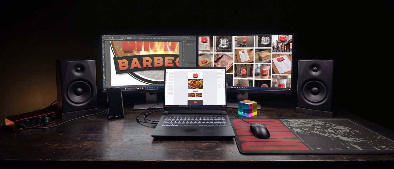 A three screen desk in a dark room, mid-job on one client's brand: the logo open in Illustrator on the left monitor, a contact sheet of that logo applied to signage, packaging and merchandise on the right, and an email campaign being built on the laptop between them. Studio speakers, an audio interface, a phone in a stand and a CMY color cube sit on the desk.