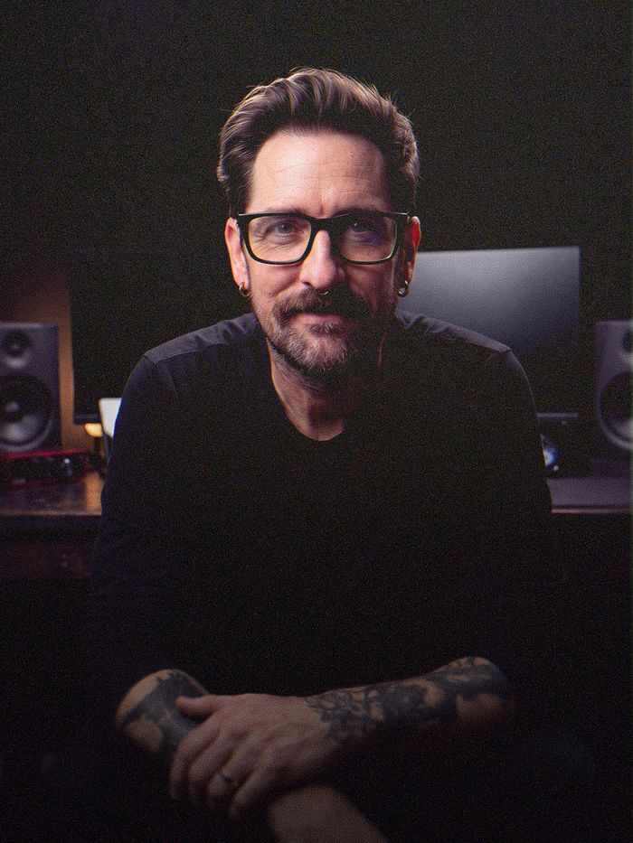 Jon May at his desk in a dark studio, seated, arms folded, wearing glasses and a black long-sleeve shirt with tattooed forearms, studio monitors and a screen behind him.