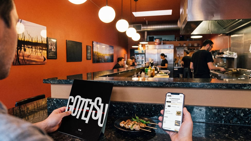 A diner at the counter holding the printed menu in one hand and the restaurant's site open on a phone in the other.