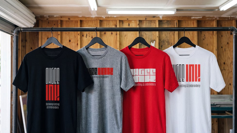 Four shirts on a shop rack in black, heather gray, red and white, each printed with a different lockup from the identity family.