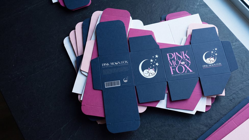 Flat printed box blanks in navy, pink and white, stacked beside a built box.