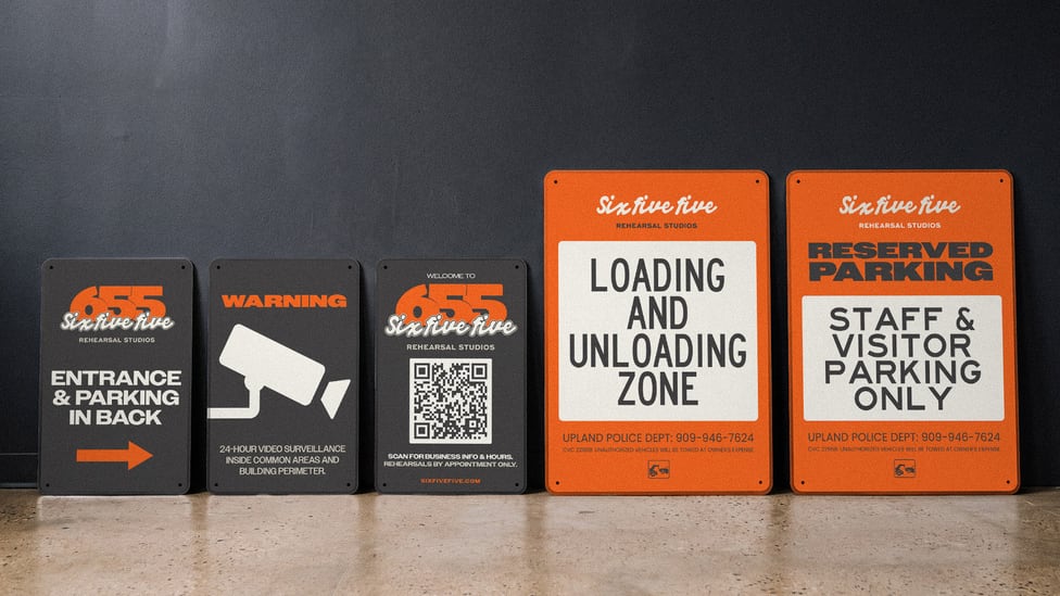 A row of 655 signs leaning against a wall: entrance direction, a video surveillance warning, a welcome sign with a QR code, a loading and unloading zone sign and a reserved parking sign.
